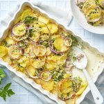 Een ovenschotel staat op tafel, waaruit al 1 hoek is uitgeschept op een bord. De ovenschotel bestaat uit krokante aardappelschijfjes, zoals een aardappelgratin, maar er is ook broccoli, rode ui en gerookte ham aan toegevoegd. Er staat een vaasje langs met een bloemetje en het tafelkleed is wit met ruitjes. 