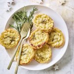 Taartjes met schijfjes aardappel en Belgische kaas liggen op een wit bord. Goud gestek ligt op het bord om meteen zo'n taartje op te scheppen. Er liggen enkele sterretjes en lichtjes langs om de feesttafel extra te laten blinken. 