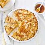 Goudgele appelcake staat op een houten witte tafel. De ronde cake is al voor een kwart aangesneden. Er is al een stukje uitgenomen en er staat een potje met abrikozengelei bij. 