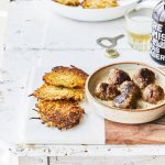 Gebakken balletjes liggen in een diep bord op de mosterdsaus, de krokante groenterösti's liggen er langs. Er staat een flesje bier op tafel met een klein glaasje.