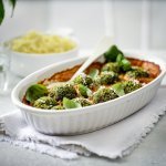 Een witte ovenschotel staat op tafel op een dikkere keukenhanddoek. De ovenschotel is gevuld met rode paprikasaus en hierop liggen de groene vegetarische balletjes van boerenkool. In de verte zie je ook nog een kommetje puree, klaar om aan tafel te gaan dus!