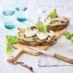 Een rijkbelegde toast met venkel en mozzarella liggen op een houten plank. Boven deze plank staan twee blauwe glazen gevuld met water. Het gerechtje is afgewerkt met enkele basilicumblaadjes. 