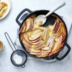 Een clafoutis van appelstukkjes staat op het aanrecht in een zwarte fonte schaal. Er is al een portie uitgeschept en staat op een bordje naar de pot. Er is ook bloemsuiker over het dessert gestrooid en de koffie is al uitgeschonken. 