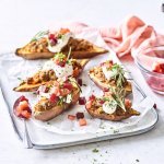 Op het aanrecht staat een rechthoekige porseleinen serveerschaal met hierop de gevulde zoete aardappelen. Het gerechtje heeft een kleur waar je blij van wordt! Het witte toefje van de zure room, blokjes rode biet en tomaat en afwerken doen we met een frisgroen takje rozemarijn. Een prachtig gerecht in smaak en kleur!