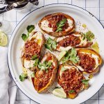 Een ronde ovenschaal met de gepofte zoete aardappelen staat op tafel. De gepofte aardappelen zijn gevuld met yoghurt en hierbovenop de chili sin carne, ze zijn afgewerkt met enkele blaadjes koriander en partjes limoen. De witte ovenschaal heeft een blauw randje en staat op een blauw geruite handdoek.