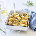 Een witte ovenschaal met donkerblauw randje staat op een keukenonderlegger op een witte houten tafel. Er staat een witte pepermolen en een glazen flesje met olijfolie langs. De verse tijm zorgt voor extra kleur bij de geroosterde aardappelen, er ligt ook nog een blauwe linnen servette langs de ovenschotel.