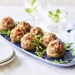 Op een blauw schaaltje liggen gevulde champignons. Ze zijn geserveerd met enkele blaadjes rucola en een stappeltje bordjes met mini-vorkjes om het hapje proper te eten. 