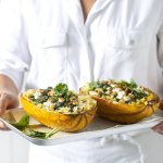 Spaghettipompoen ligt doormidden gesneden op een ovenbakplaat, gevuld met spinazie en kaas. De vrouw heeft de ovenschaal vast en draagt een wit hemd met witte schort. 