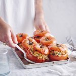Zes gevulde rode tomaten in een ovenschaal zijn aan tafel gebracht door een vrouw in witte t-shirt. Ze schept alvast eentje op voor zichzelf.