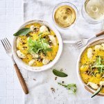 Gnocchi met pompoensaus en salie staan klaar op een tafel met wit tafelkleed. Bestek met houten afwerking ligt bij het bord, het gerechtje is afgewerkt met verse blaadjes salie en pitten. 