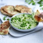 Krokant gebakken platbrood ligt op een serveerschaaltje waarbij een dip van erwten en roomkaas in geserveerd. De dip is gemixt en heeft een mooie groene kleur, om af te werken zijn er nog enkele verse erwtjes bovenop gestrooid. Een hand neem alvast een stukje platbrood om te dippen in gezonde groentedip. 