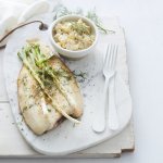 De gebakken vis ligt op een stenen witte snijplank, waar de gigrilde lente-ui bovenop ligt. De Macédoine van venkel en krieltjes is opgediend in een apart wit potje. Er ligt wit bestek langs, met een glaasje witte wijn en een linnen servette. Het gerechtje is afgewerkt met verse dille.