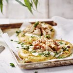 Twee naanbroodjes liggen op een metalen ovenplaat. Ze zijn belegd met yoghurt, stukjes gebakken kip en afgewerkt met salsa verde. De ovenplaat staat op een witte houten tafel.