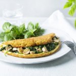 Een heerlijke dichtgevouwde omeletsoufflé gevuld met champignons, spinazie en kaas ligt op een wit bord. Het gerechtje werd afgewerkt met een slaatje van verse kruiden, denk maar aan basilicum en dille.  