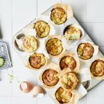 Ontbijtmuffins met courgette, yoghurt en Belgische kaas in een grote muffinvorm. Klaar om eruti te halen en op te eten. Naast de bakvorm ligt een verse courgette en lege eierschalen voor de sfeer.