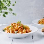 Een portie pasta met homemade paprikasaus staat klaar op een witte houten tafel. Een vers plantje oregano staat achteraan in beeld. 