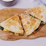 Een klaargemaakte quesadilla ligt op een houten snijplank. De quesadilla is in 3 gesneden, hij is gevuld met vlees, spinazie, zwarte bonen en kruiden. 