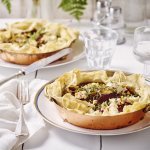 Aan tafel! Quiche van filodeeg met geitenkaas en rode biet staan klaar op tafel. De quiches zitten in een kopperen schaaltje en staan op een wit bord. Het bestek en de witte servette liggen langs het bord. Op tafel staan ook waterglazen en een glazen peper- & zoutvat.