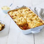 Een lichtblauwe ovenschotel staat op een houten witte tafel. De ovenschotel is gevuld met een mac & cheese én een tomatensaus, waardoor hij eerder weg heeft van een lasagne. Er is al 1 portie uitgeschept en wil al beginnen aan de volgende. Laat het niet koud worden. 