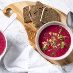 Soep van rode biet kleurt natuurlijk rood en smaakt naar nog! Ze is geserveerd in twee witte kommetjes en staat op een houten snijplank met enkele bruine sneden brood bij. 