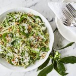 Een gezonde side dish voor je feestgerecht dat extra stukje power te geven. Deze spruitjessalade is opgediend in een witte keramische kom met stukjes appel en afgewerkt met een dressing op basis van blauwekaas, zure room en mayonaise. Er ligt bestek langs op een witte nap. 