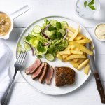 Een echte Vlaamse klassieker die smaakt voor jong & oud, een steak mooi gebakken met heerlijke goude frietjes en een salade. De champignonsaus staat langs het bord in een sauspotje. Bestek en servette liggen langs het bord klaar om te starten. 