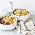 Twee witte stoofpotjes sttaan op een witte houten tafel en zijn gevuld met traag gegaard rundsvlees. De stoofpot is afgedekt met een laagje van aardappelschijfjes. De gratin is mooi goudgeel gebakken. Er staat een pepermolen langsheen de kookpotten, servette en grote lepel.