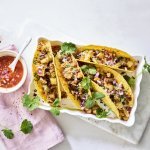 Vier tacos liggen klaar in een ovenschaal om aan te vallen. De pittige tomatendip staat links van de schotel op een roze servette. Het gerechtje is afgewerkt met verse korianderblaadjes.