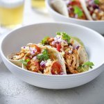 In een diep wit bord liggen twee gevulde taco's met pulled chicken, afgewerkt met tomaat, rode ui en geraspte Passendael kaas. Om wat extra kleur te geven liggen er enkele blaadjes koriander op. Wij serveren ze met frisgekoeld biertje, smakelijk!