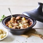 Zwartkleurige tajine met gehaktballetjes van lamsvlees, afgewerkt met verbrokkelde feta. Er staat een portie couscous naast de tajine. 