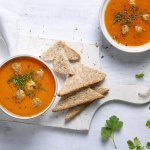 Heerlijk gemaakte tomatensoep staat uitgeschenkt op tafel in twee soepborden. De balletjes drijven in de soep en is afgewerkt met peterselie. Brood en lepels liggen langs het bord, klaar om te slurpen.