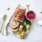 Een mooi aangesneden varkensrollade ligt op een rechthoekig dienblad. De aardappeltjes werden net niet doormidden gesneden alvorens ze de oven in gingen met een kruidentakje. De veenbessencompote staat langs de rollade die zorgt voor een intense kleur in deze feestmaaltijd. Goud bestek ligt langs het gerecht. 