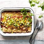 Enchiladas liggen mooi op een rijtje in een witte ovenschotel. Hierop werd tomatensaus geserveerd en gemalen kaas, in de oven. Versgesneden lente-uitjes werden als afwerking toegevoegd aan het gerecht. 