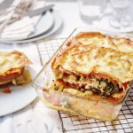 De lasagne staat op tafel in een glazen doorzichtige ovenschotel. Hij is aangesneden zodat je mooi de laagjes ziet van pasta, groenten en saus. De portie die is uitgeschept ligt op een bordje er naast.