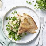 Een wit bord gevuld met een frisse schep van de puree boordevol extra groentjes, denk maar aan verse lente-uitjes, zongedroogde tomaatjes, waterkers, kruiden enz. Hierop werd de gebakken rogvleugel gelegd die mooi krokant is gebakken. Naast het bord ligt ook nog een lichtblauwe servette, kommetje met extra waterkers en een glaasje wijn. Smakelijk!