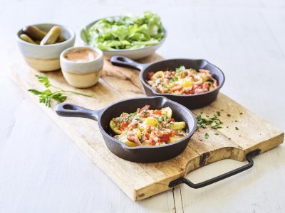 Aardappelplakjes met spek, paprika en kaas voor de gourmet Twee gourmetpannetjes staan te sieren op een houten snijplank. Als bijgerechtjes hebben we een groene salade, opgelegde augurkjes en een cocktailsausje.