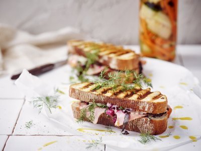 Croque met ham, snijbiet en Brugge kaas Twee croques liggen op het aanrecht in de keuken op een wit boterpapiertje. In de achtergrond staan de gepekelde groentjes (wortel en komkommer) in een bokaal. De tosti's zijn mooi gegrild in streepjes.