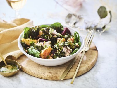 Een salade van bietjes, boerenkool en spinazie afgewerkt met noten en makreel. Deze feestsalade staat op een houten plank met de dressing in een apart gouden potje.