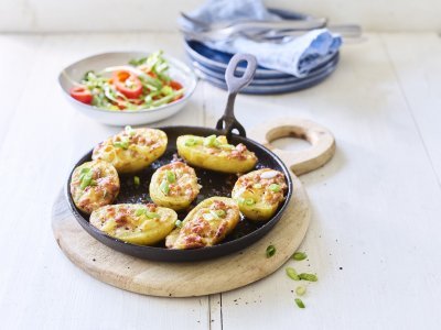 Gevulde aardappelen met spek en Flandrien kaas op de barbecue 7 gevulde aardappelen liggen op een zwarte vuurvaste pan, ze zijn gevuld met aardappelkruim, kaas en spekblokjes. Er ligt een handdoek achteraan in beeld en een slaatje met tomaatjes.