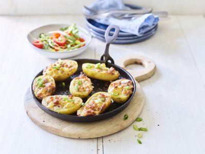 7 gevulde aardappelen liggen op een zwarte vuurvaste pan, ze zijn gevuld met aardappelkruim, kaas en spekblokjes. Er ligt een handdoek achteraan in beeld en een slaatje met tomaatjes. 