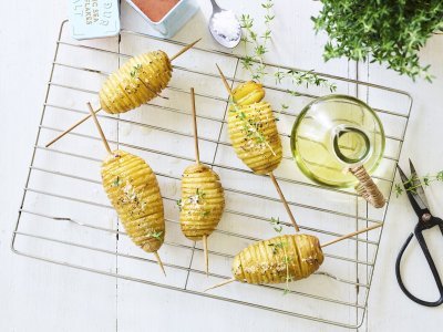 Hasselback potatoes liggen op een bakrooster te blinken. Ze zijn doorprikt met lange houten prikker om makkelijker te kunnen draaien op de BBQ. Er staat een flesje olie, verse tijm en een potje zeezout naast. 