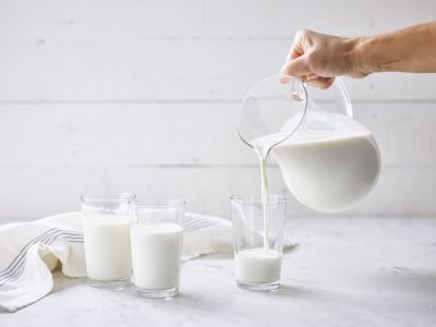 Glazen melk worden gevuld 2 glazen zijn al gevuld met melk. Glazen kan met melk vult nog een glas. Glazen melk staan op marmere keukenaanrecht met linnen handdoek.
