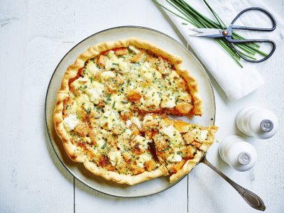 Quiche of groententaart van zoete aardappel met verse geitenkaas. Aangesneden op een wit bord met zwart randje en zilveren taartschep. Links van het bord staan witte molens van peper en zout en rechtsboven liggen enkele bieslooktakjes met keukenschaartje. Dit alles op een witte houten tafel.