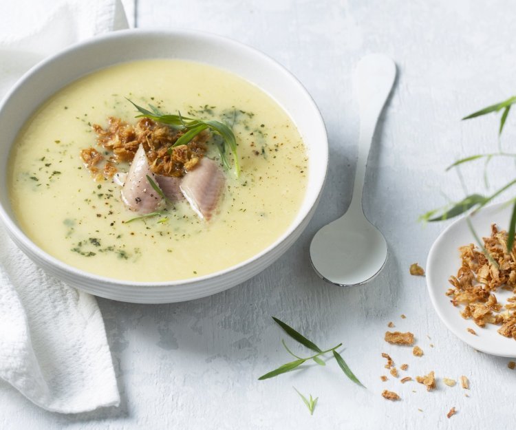 Aardappelsoep met gerookte paling en krokante uitjes Dampende aardappelsoep met stukjes paling in het midden. We geven ze extra kleur en smaak door dragon toe te voegen en krokante uitjes. Langs het bord ligt een witte eetlepel en extra uitjes en dragon.