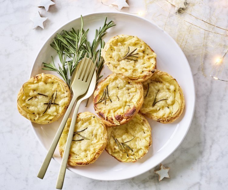 Taartjes met schijfjes aardappel en Belgische kaas liggen op een wit bord. Goud gestek ligt op het bord om meteen zo'n taartje op te scheppen. Er liggen enkele sterretjes en lichtjes langs om de feesttafel extra te laten blinken. 