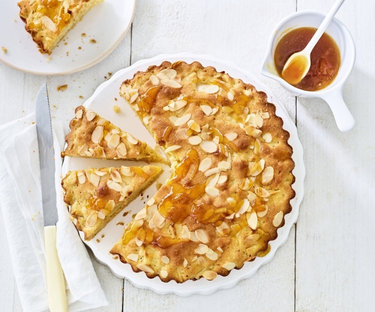 Appelcake met amandelen Goudgele appelcake staat op een houten witte tafel. De ronde cake is al voor een kwart aangesneden. Er is al een stukje uitgenomen en er staat een potje met abrikozengelei bij.