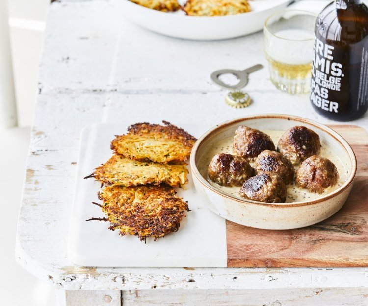 Balletjes in mosterdsaus met groenterösti Gebakken balletjes liggen in een diep bord op de mosterdsaus, de krokante groenterösti's liggen er langs. Er staat een flesje bier op tafel met een klein glaasje.