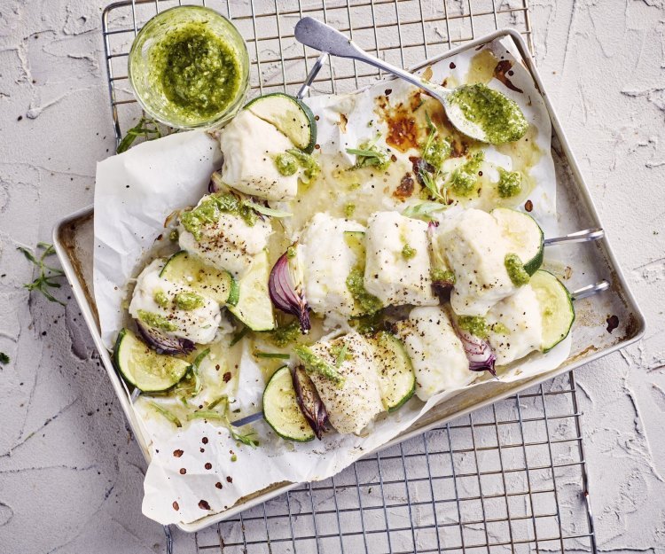 Brochette van heek met courgette, rode ui en dragonpesto Klaargemaakte brochettes liggen in een grijze ovenschaal te blinken. De spiesjes bestaan uit stukjes heek, courgette en rode ui. Er staat een glazen potje naast met huisgemaakte dragonpesto.