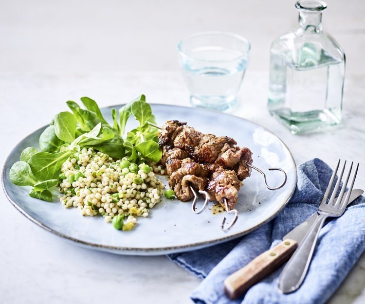 Brochette van rundvlees met salade van parelcouscous, erwtjes, verse kruiden en veldsla Op een lichtblauw groot bord liggen 3 gegrilde brochettes, we serveren ze met een couscoussalade van erwtjes en verse kruiden. Naast het bord ligt een blauwe servette met bestek langs. Er staat ook een glaasje water bovenaan het bord met een flesje water naast.