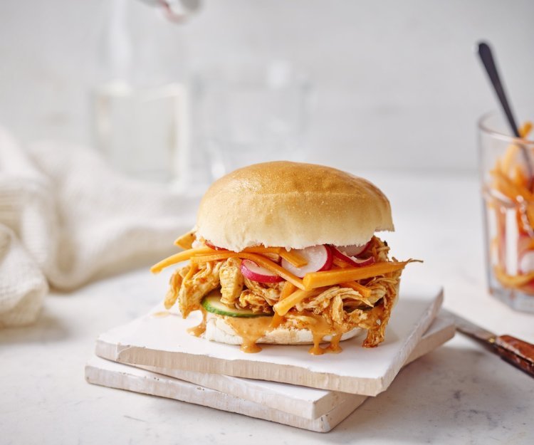 Broodje pulled chicken met chilimayonaise Een heerlijke pistolet ligt op een houten snijplankje. Het is opengesneden en gevuld met de boterzachte kip, zoetzure groentjes en chilimayonaise. In de achtergrond ligt nog een servette om je smosvlekken snel op te drogen.